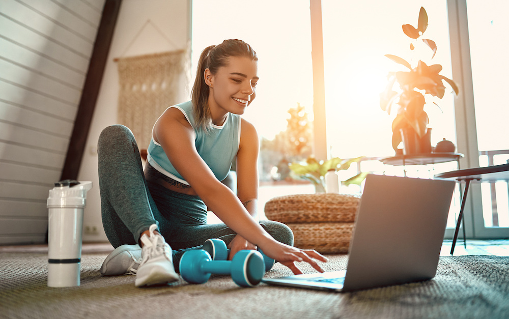A sporty woman in sportswear is sitting on the floor with dumbbells and a protein shake or a bottle of water and is using a laptop at home in the living room. Sport and recreation concept. Fitness leads
