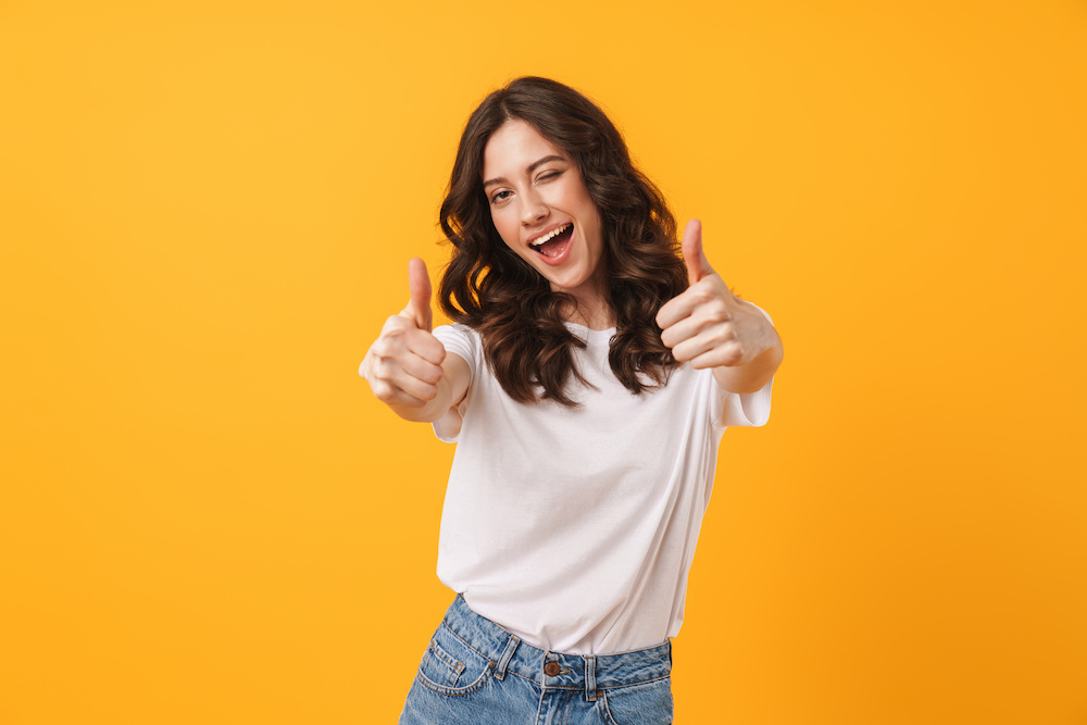 Image of beautiful brunette woman wearing casual clothes winking and showing thumbs up at camera